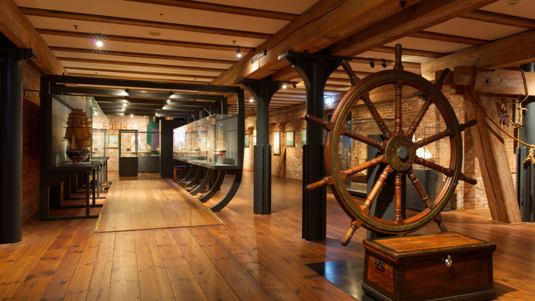 Großes antikes hölzernes Steuerrad auf einer Truhe im Vordergrund eines maritimen Museums mit warmem Holzfußboden, sichtbaren Deckenbalken, Backsteinwänden und Glasvitrinen mit Schiffsmodellen.