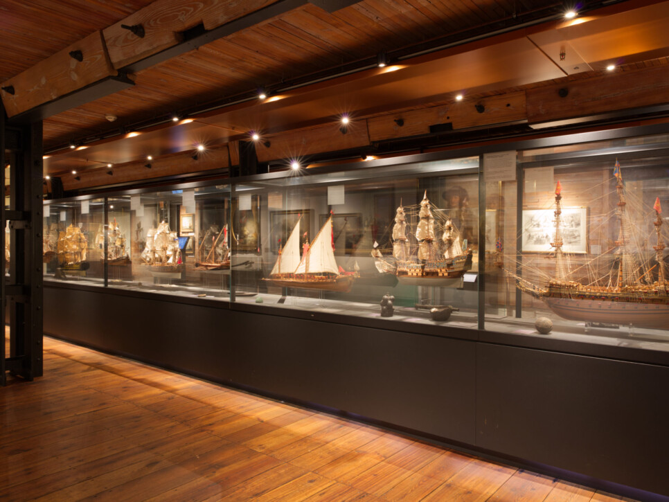 Innenansicht eines maritimen Museums mit einer langen Glasvitrine, die mehrere detaillierte historische Schiffsmodelle zeigt; warmer Holzboden, sichtbare Deckenbalken und Spotbeleuchtung.