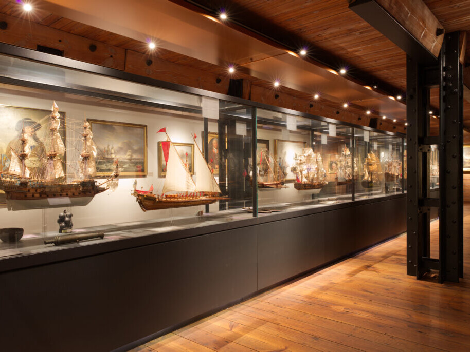 Langgestreckte Glasvitrine mit detaillierten historischen Schiffsmodellen und maritimen Gemälden in einem warm beleuchteten Museumsraum mit Holzdecke, Stahlträgern und Parkettboden.
