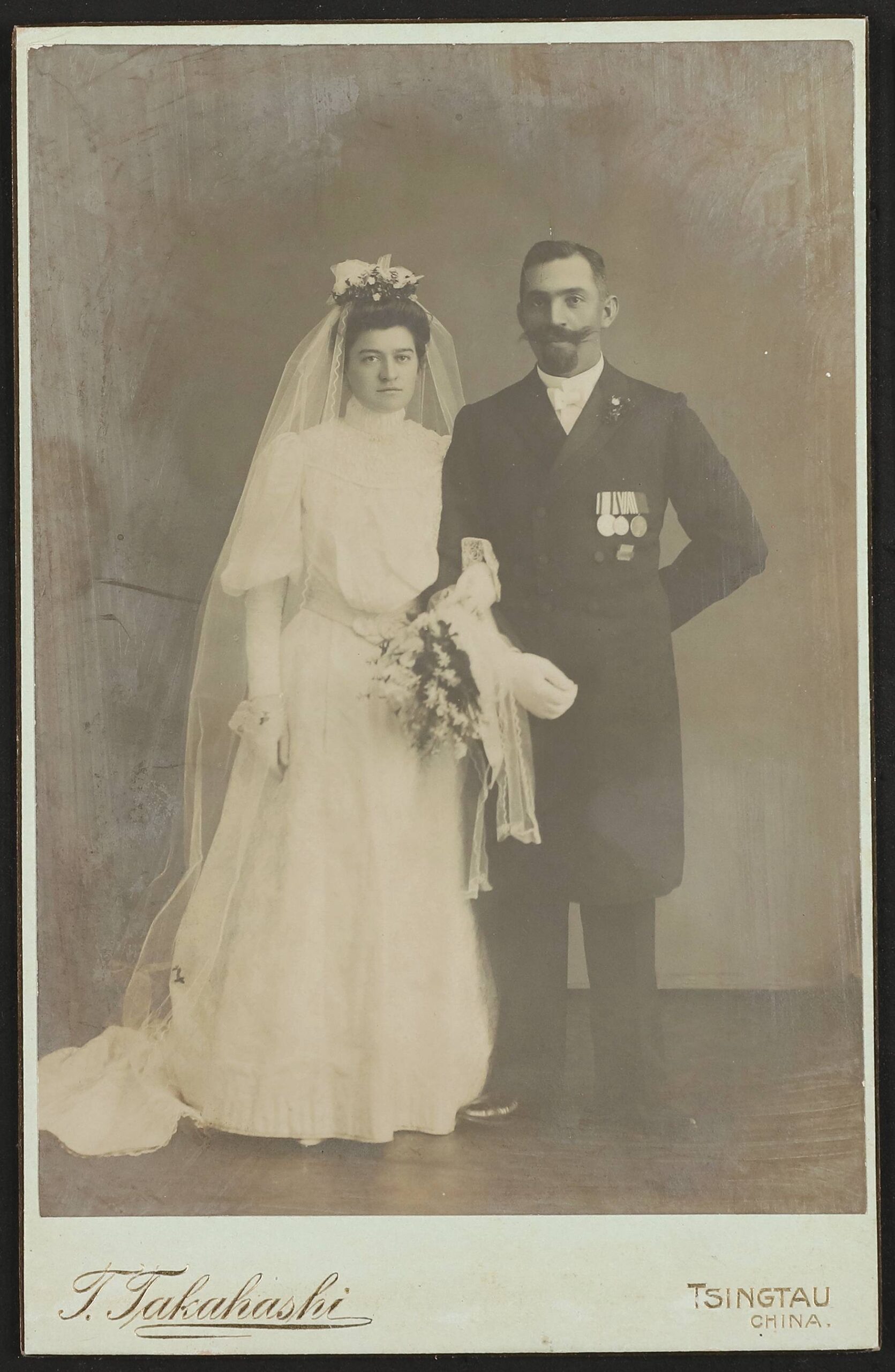 Porträt eines Brautpaars: Frau in weißem Kleid mit Schleier und Strauß, Mann mit Schnurrbart, Anzug und Medaillen; Aufdruck T. Takahashi Tsingtau