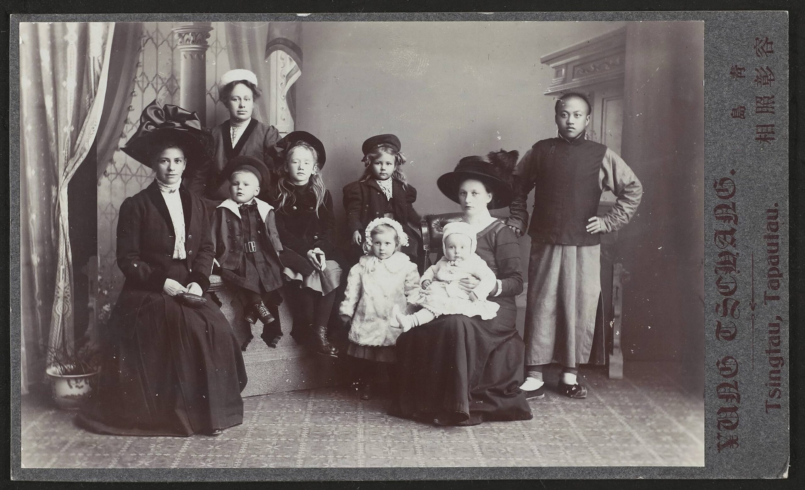 Neun Personen in einem Studio-Porträt: Frauen und Kinder in historischer Kleidung mit großen Hüten, rechts ein Mann in dunkler Jacke und weitem Rock.