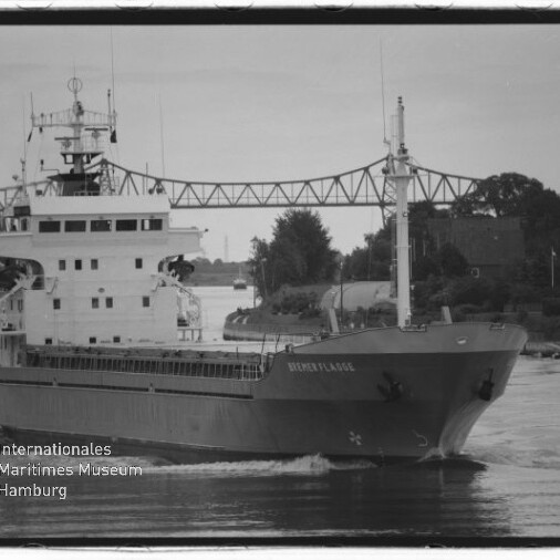 Frachtschiff fährt einen Fluss entlang unter einer Stahlbrücke, schwarzweißaufnahme mit Museumslogo in der Ecke.