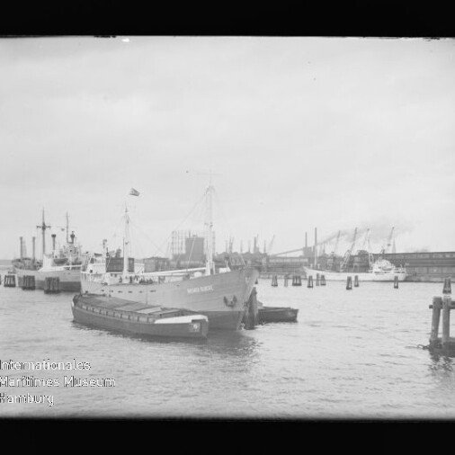 Frachtschiff mit Beiboot im Hafen vor Kaimauern und Kränen; links unten Wasserzeichen 