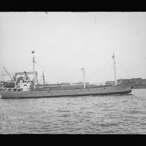 Frachtschiff mit zwei Masten und Ladekränen auf ruhigem Wasser, Hafenbebauung im Hintergrund.