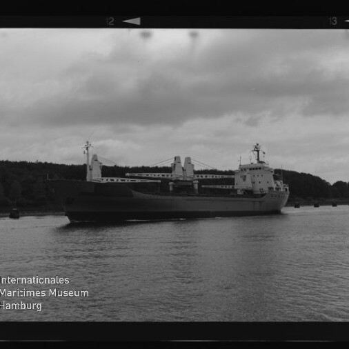 Schwarzweißfoto eines Frachtschiffs mit Ladekränen vor bewaldeter Küste; Text: Internationales Maritimes Museum