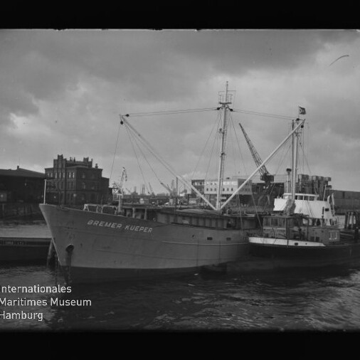 Frachtschiff Bremer Kueper am Kai mit Ladebaum-Masten und Schute daneben, Hafenbauten im Hintergrund.