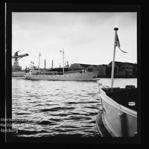 Schwarzweiß-Hafenbild: großes Schiff mittig, kleines Boot mit Flagge vorn, Werft hinten, Wasserzeichen Maritimes Museum