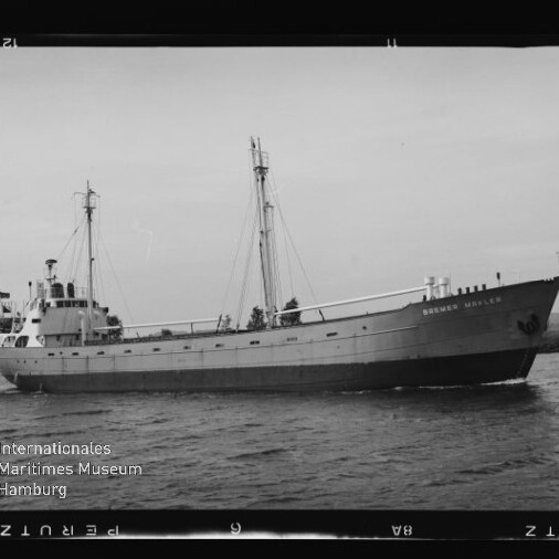 Graues Schwarzweißfoto eines Frachtschiffs mit zwei Masten und sichtbarer Aufschrift am Bug, fahrend auf einem Kanal vor baumbewachsenem Ufer.