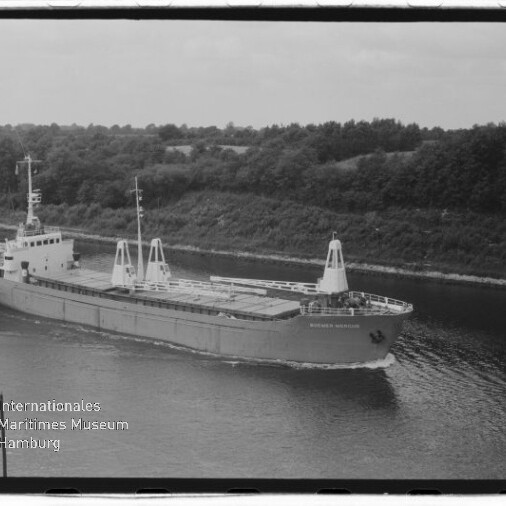 Frachtschiff mit Ladebühnen und Aufbauten fährt durch einen Kanal vor baumbewachsenem Ufer; Museumslogo unten links.