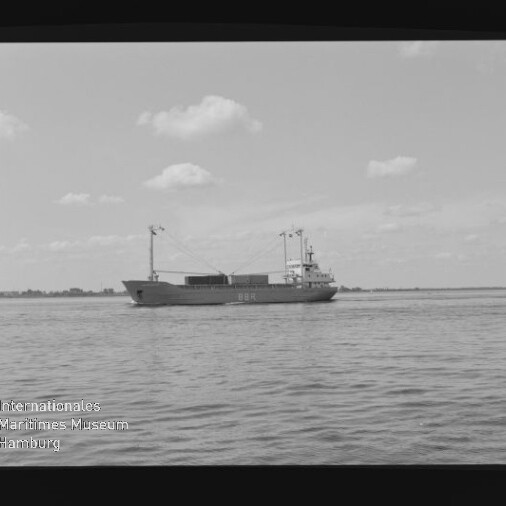 Schwarzweißfoto eines Frachtschiffs auf ruhiger See unter leicht bewölktem Himmel; Filmrand und Museumswasserzeichen sichtbar.