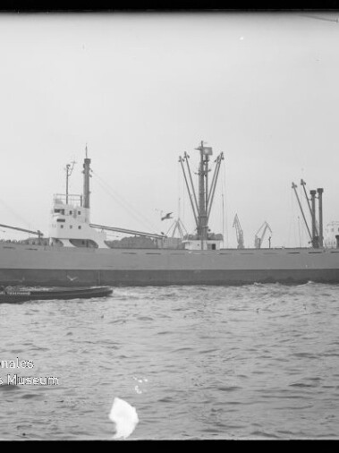 Frachtschiff mit mehreren Ladekränen und Masten, kleines Begleitboot im Vordergrund und Hafen im Hintergrund, Schwarzweißfoto