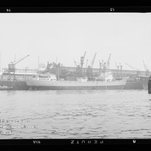 Schiff am Industriehafen an der Kaimauer, mehrere Hafenkräne im Hintergrund und Wasser im Vordergrund.