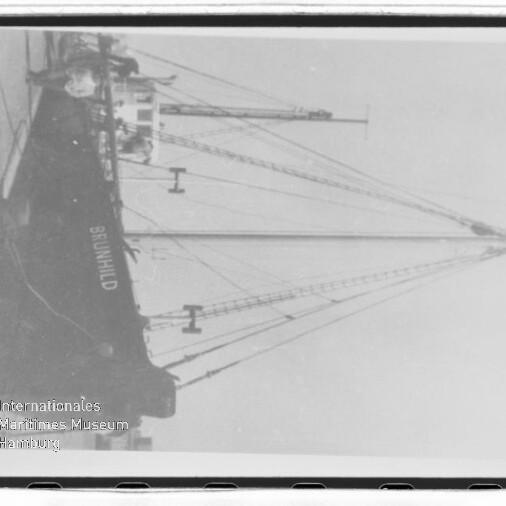 Schwarzweißfoto eines am Kai liegenden Frachtschiffs mit Masten, Name BRUNHILD und Wasserzeichen des Maritimen Museums Hamburg.