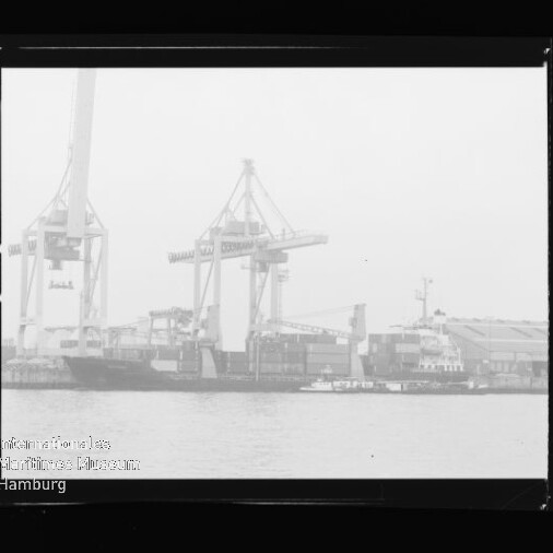 Frachtschiff unter großen Hafenkränen an der Kaimauer, kleines Schleppboot davor; Rahmen mit Maritimes Museum Hamburg.