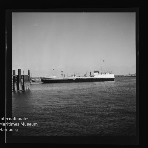 Schwarzweißaufnahme eines größeren Frachtschiffs vor einem Pier im Hafen, Museumslogo 