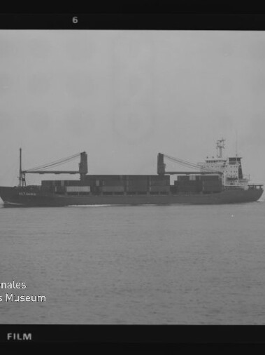 Schwarzweißaufnahme eines Frachtschiffs in Seitenansicht auf ruhiger See mit zwei Ladekränen und Containerstapel, Museumslogo unten links.
