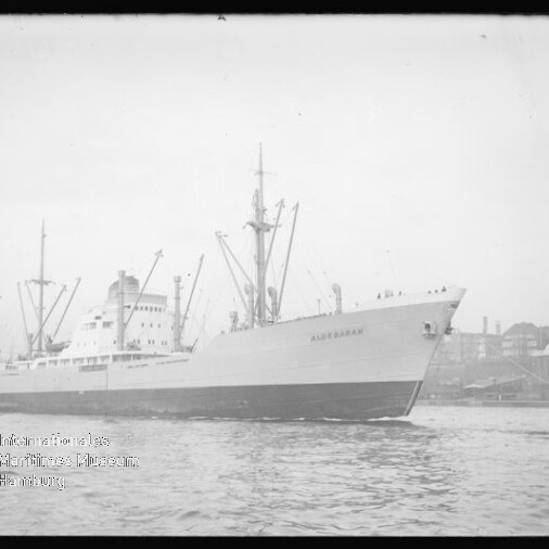 Großes Frachtschiff mit Ladekränen und sichtbarem Schriftzug am Bug fährt in einen Hafen, im Hintergrund Industrieanlagen.