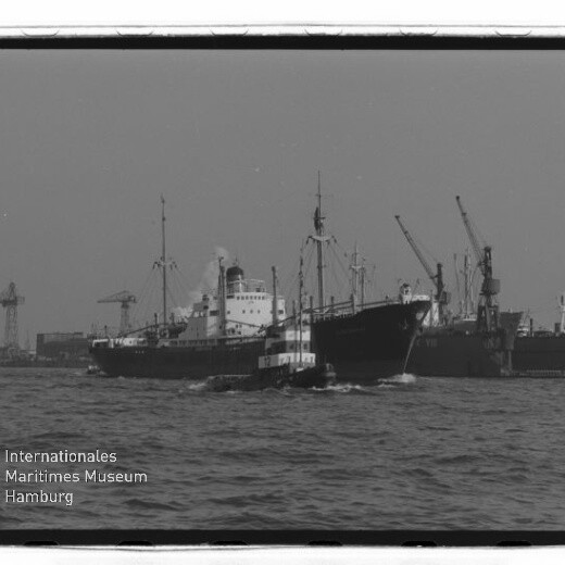 Schlepper begleitet großes Frachtschiff vor Hafenkränen, mit Schriftzug Internationales Maritimes Museum Hamburg unten links.