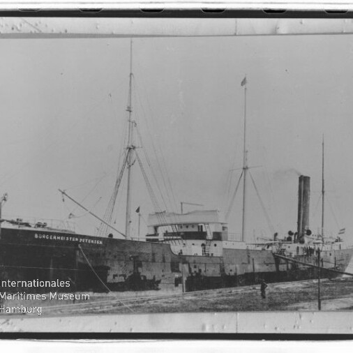 Historisches Dampfschiff Bürgermeister Petersen mit Schornstein und hohen Masten am Ufer, schwarzweiß.