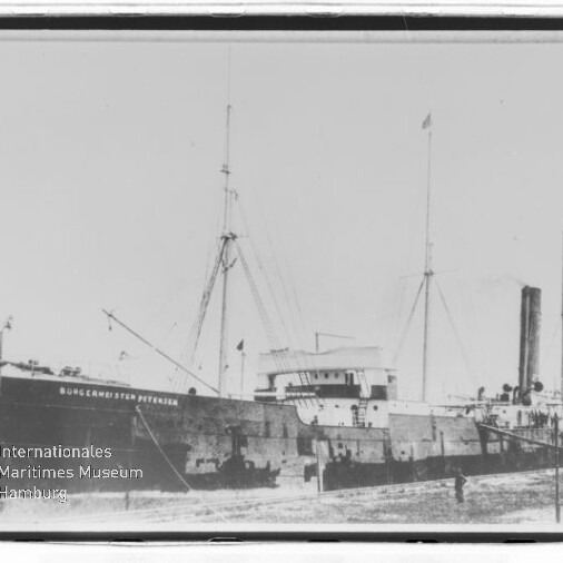 Frachtschiff mit Schornstein und Masten am Kai, Rumpf beschriftet und Wasserzeichen des Internationalen Maritimen Museums Hamburg.