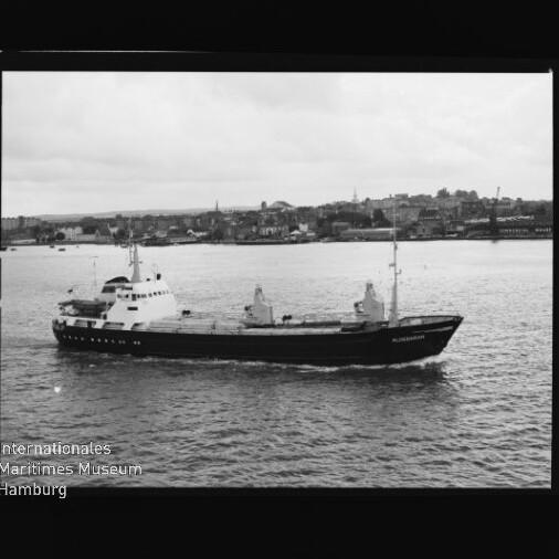 Schwarzweißbild eines Frachtschiffs namens Alexandria, das vor der Uferlinie einer Hafenstadt mit Kirchtürmen fährt.