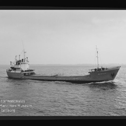 Kleines Frachtschiff mit Aufbauten achtern und Mast vorn auf ruhiger See; Schwarzweißfoto mit Wasserzeichen unten links.