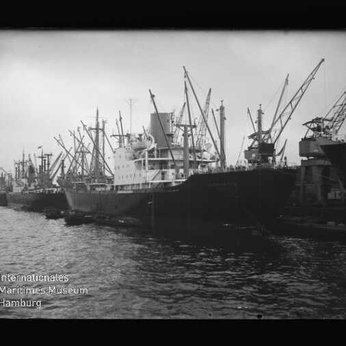 Mehrere Frachtschiffe mit Kränen an einem Hafenpier, Wasser im Vordergrund.