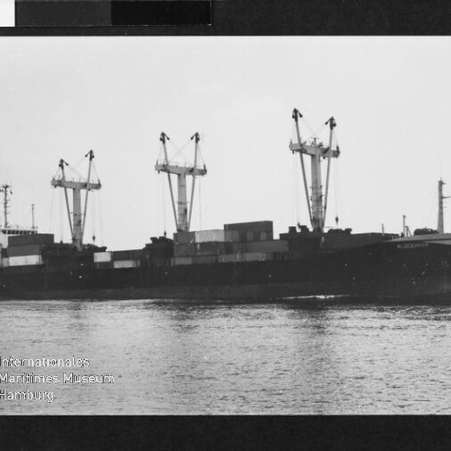 Schwarzweißfoto eines Frachtschiffs mit drei Ladekränen und Containern auf ruhigem Wasser, Museumsstempel unten links.