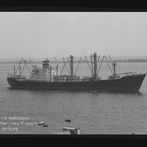 Frachtschiff mit mehreren Ladekränen und mittschiffs gelegenem Aufbau auf ruhigem Meer, Küstenlinie im Hintergrund.