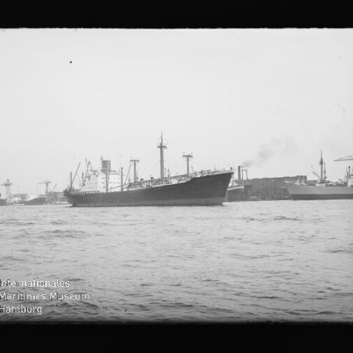 Frachtschiff mit Schornstein und Ladekränen im Hafen, Kaimauern und Kräne im Hintergrund; Wasserzeichen unten links.