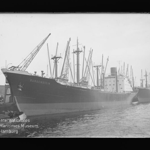 Großes Frachtschiff längsseits am Kai mit mehreren Ladekränen und Masten, schwarz-weiß aufgenommen.