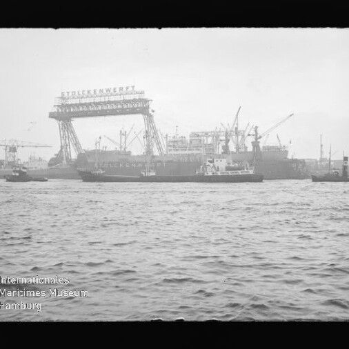 Blick über das Wasser auf eine Werft mit großem Gitterrahmenkran, mehreren Frachtschiffen und zahlreichen Hafenkränen.