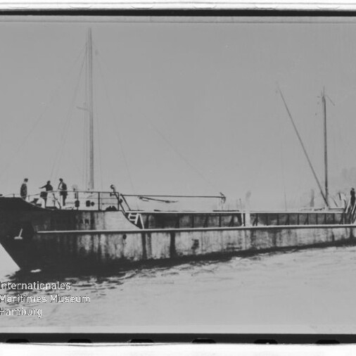 Flaches Frachtschiff mit zwei Masten und mehreren Personen auf dem Deck, fahrend auf dem Wasser.