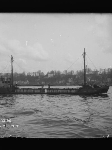 Frachtschiff mit zwei Masten auf ruhigem Wasser vor Ufer mit Häusern; Wasserzeichen des Int. Maritimen Museums Hamburg.