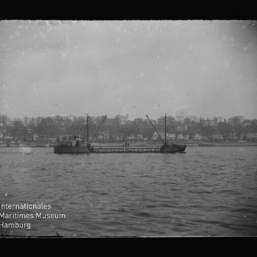 Frachtschiff mit zwei Ladekränen auf ruhigem Wasser, Häuser und Bäume am Ufer im Hintergrund; Museumslogo unten links.
