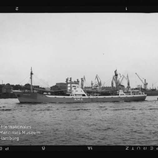 Frachtschiff mit HMG am Rumpf fährt durch einen Industriehafen mit Kränen, daneben ein kleines Schlepperboot.