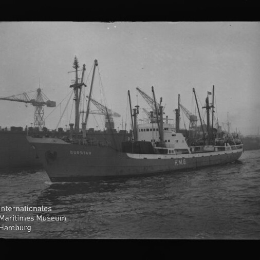 Frachtschiff mit sichtbaren Buchstaben HMG an Bord fährt im Hafen vorbei, im Hintergrund mehrere Hafenkräne.