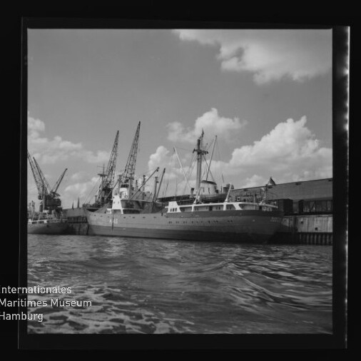 Schiff am Kai mit Hafenkränen, welliges Wasser im Vordergrund und Museumslogo 