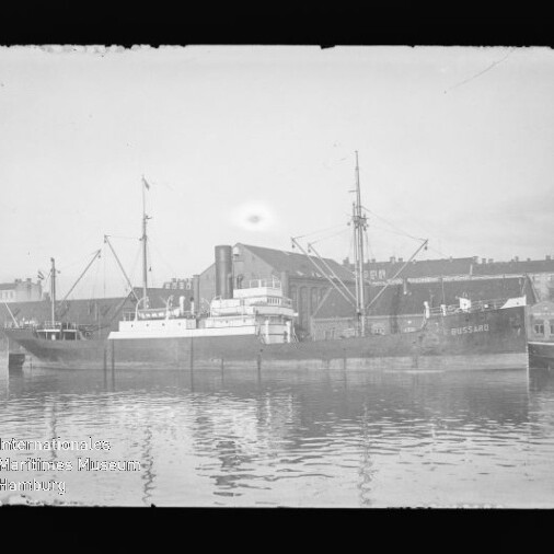 Schwarzweiß-Foto eines Frachtschiffs mit Masten und Aufbauten im Hafen vor Industriegebäuden, Spiegelung im Wasser.