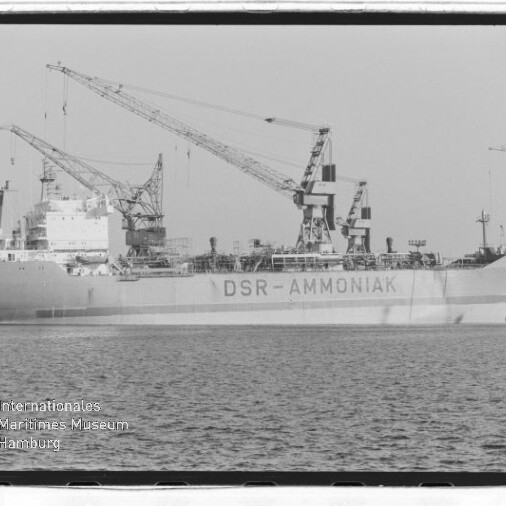Großes Tankschiff mit Aufschrift DSR‑AMMONIAK am Hafenpier, mehrere Ladekräne darüber und ruhige Wasserfläche.