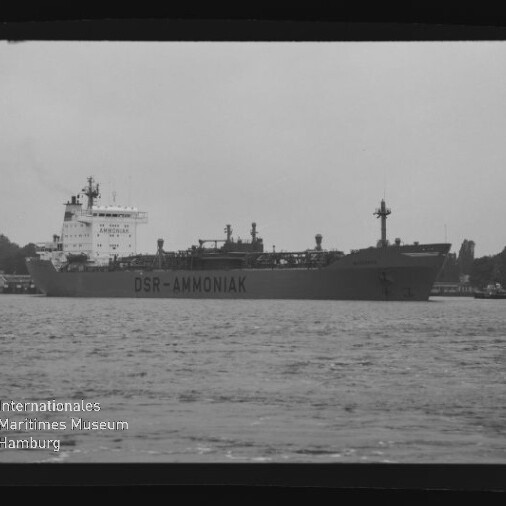Schwarzweißaufnahme eines Frachttankers mit Aufschrift DSR-AMMONIAK auf dem Wasser vor bewaldetem Ufer.