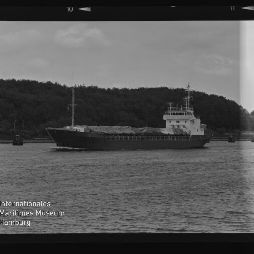 Frachtschiff auf ruhigem Wasser vor bewaldeter Küste, Schwarzweißfoto mit Museumslogo Internationales Maritimes Museum Hamburg.