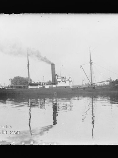 Schwarzweißaufnahme eines dampfgetriebenen Frachtschiffs mit rauchendem Schornstein und Spiegelung auf ruhigem Wasser.