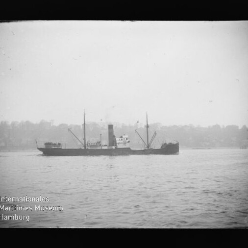 Frachtschiff mit Schornstein und Ladekränen auf ruhigem Wasser, Ufer im Dunst im Hintergrund.