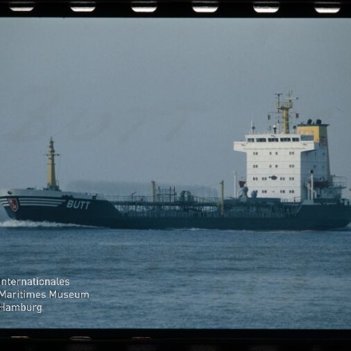 Tanker mit weißen Aufbauten und dunkelblauem Rumpf mit dem Namen BUTT fährt über ruhige See; Museumslogo unten links.