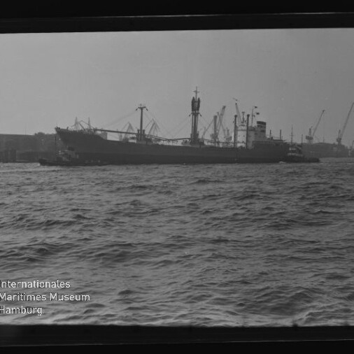Frachtschiff am Hafen mit Kränen und welliger See im Vordergrund; sichtbares Wasserzeichen 'Internationales Maritimes Museum'.