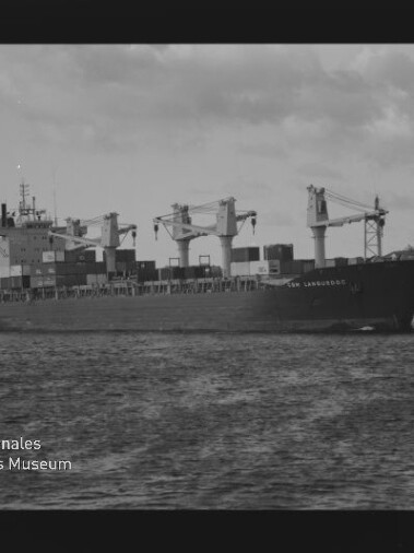 Frachtschiff mit drei Deckskranen und gestapelten Containern auf ruhigem Wasser, Küstenlinie im Hintergrund.