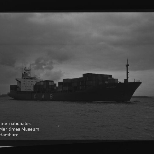 Containerschiff mit aufgestapelten Containerkästen und CMB-Schrift auf dem Rumpf fährt bei dunklem, bewölktem Himmel auf dem Meer; Museumslogo und Filmrand sichtbar.