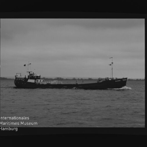 Kleines Frachtschiff mit Flaggen fährt auf ruhiger See, schwarzweiß-Aufnahme mit Rand und Museumslogo unten links