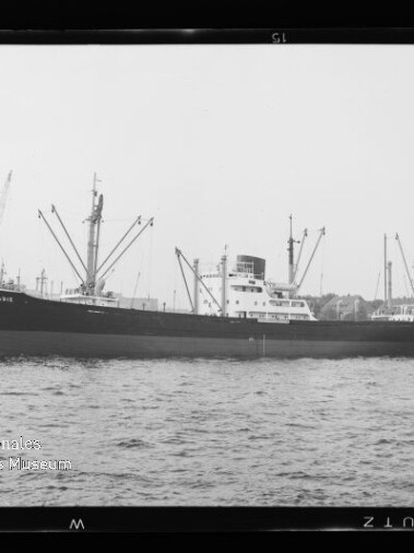 Schwarzweiß-Seitenansicht eines Frachtschiffs im Hafen vor Kränen und ruhiger Wasserfläche.